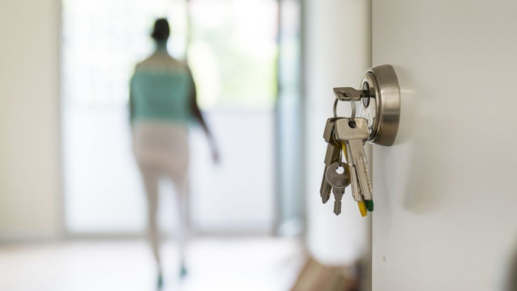 Unas llaves dentro de la cerradura de una puerta y de fondo una mujer en el interior de un piso