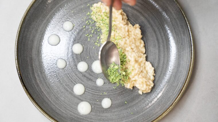 Emplatat de l'arròs cremós amb coliflor amb unes gotetes de crema de coliflor a una banda del plat i la cuinera va posant bròcoli esmicolat per sobre