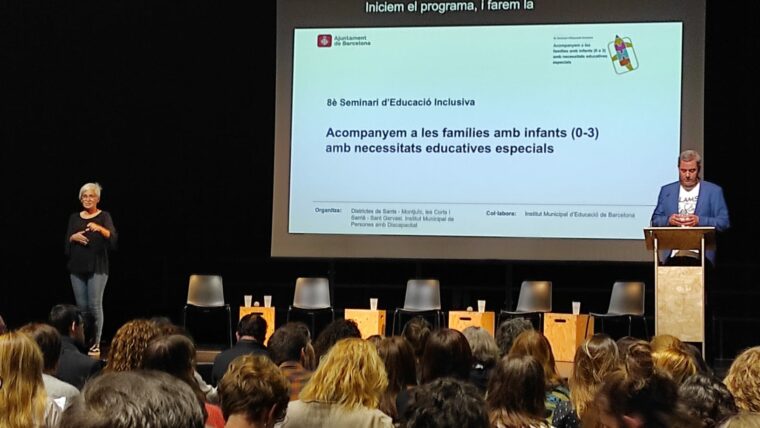 Un home i una intèrpret en llengua de signes intervenen en el seminari davant un auditori ple. A la pantalla: 8è Seminari d'Educació Inclusiva: Acompanyem les famílies amb infants (0-3) amb necessitats educatives especials.