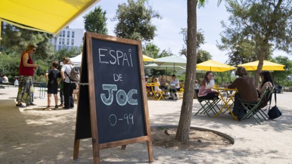 Detall d’una pissarra amb “Espai de joc 0-99” i de fons grups de famílies i joves gaudint del servei.