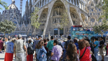 Turistes a la plaça de la Sagrada Família davant de la façana de la Passió del Temple Expiatori de la Sagrada Família amb un bus turístic passant pel carrer de Sardenya.
