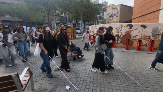 Diverses alumnes d'arquitectura passegen pel carrer fent ús d'un bastó per a persones cegues i amb els ulls tapats amb un antifaç. Totes elles van acompanyades d'una persona que les guia.