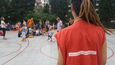 Una monitora, d&#039;esquenes, en un pati escolar obert, mentre famílies juguen.