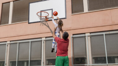 Un pare aixeca en braços a la seva filla per ajudar-la a encistellar una pilota de bàsquet.