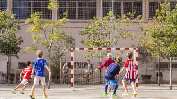 Un grup d'infants i adults juguen a futbol en un pati escolar obert durant les vacances.